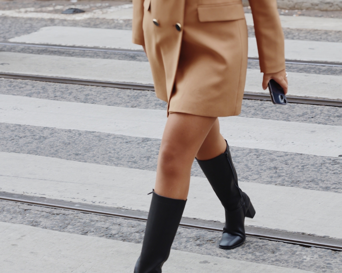 Mujer elegante con blazer y botas caminando por una animada calle urbana.