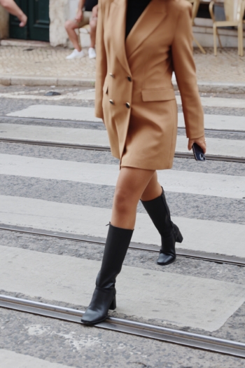 Mujer elegante con blazer y botas caminando por una animada calle urbana.