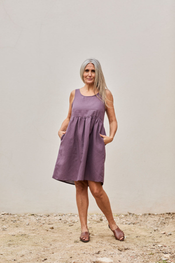 Mujer sonriente con vestido morado elegante, posando al aire libre con un fondo minimalista.