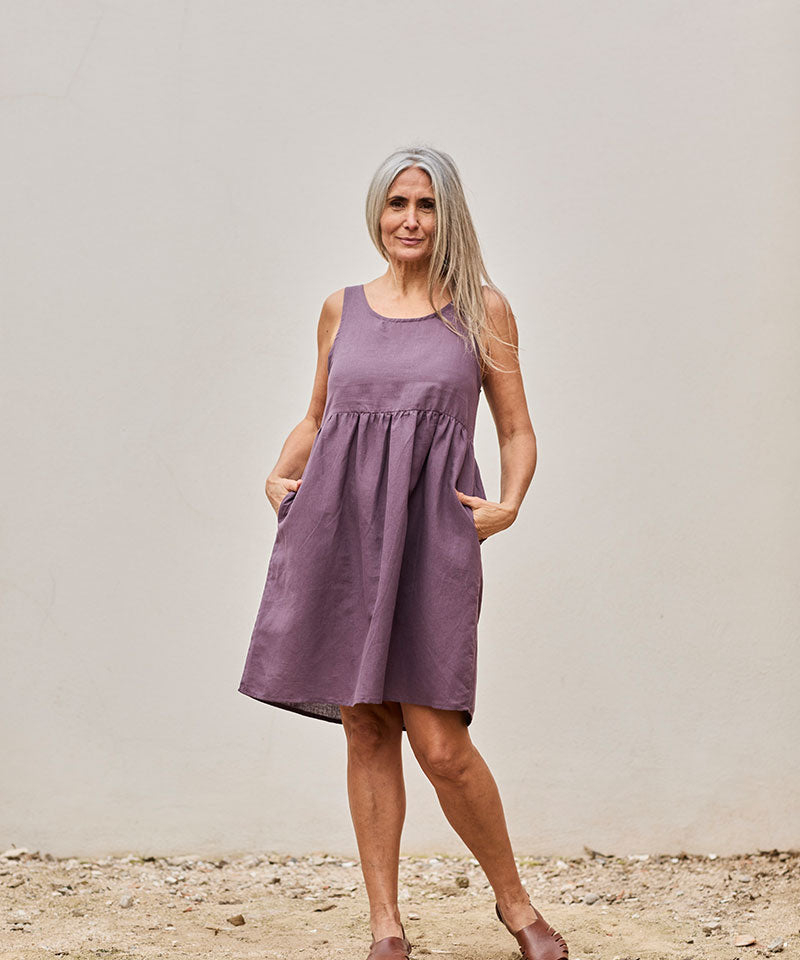 Mujer sonriente con vestido morado elegante, posando al aire libre con un fondo minimalista.