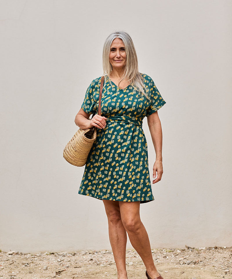 Mujer sonriente con vestido vibrante y bolso de paja en un fondo neutro.