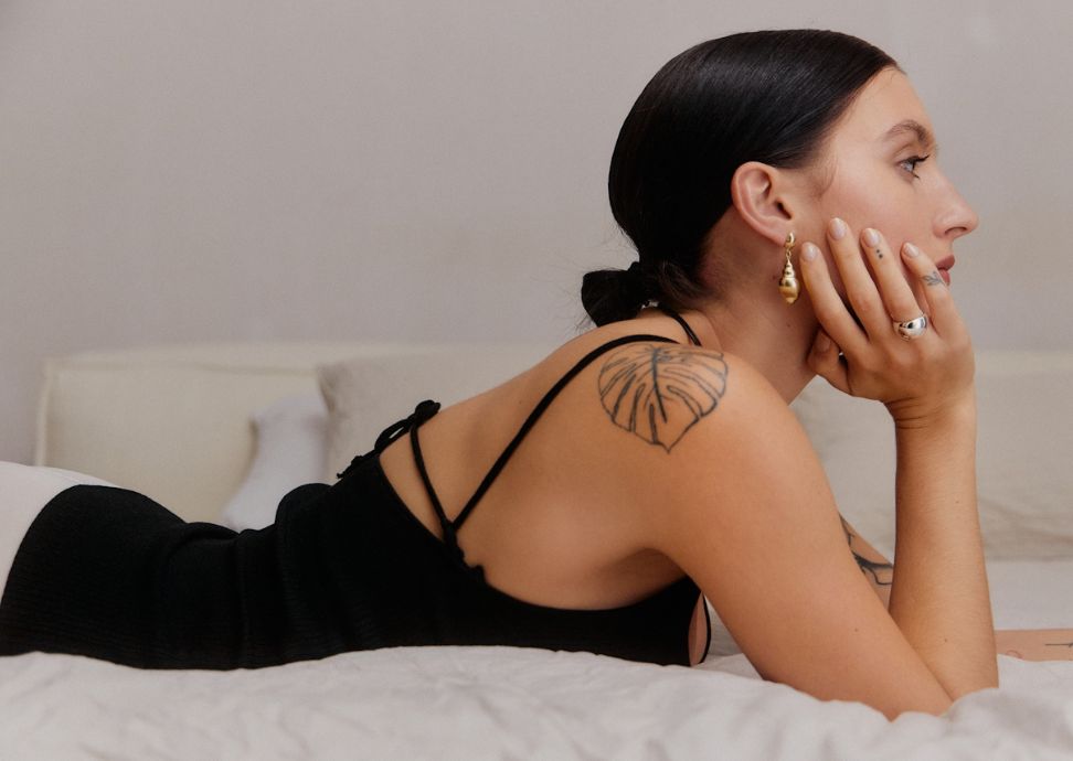 Mujer reflexionando en la cama, destacando su elegante atuendo y tatuaje de hoja.