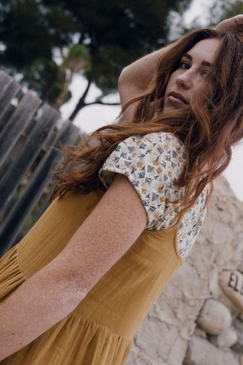 Joven mujer con vestido mostaza en un entorno natural, mirando pensativa sobre su hombro.