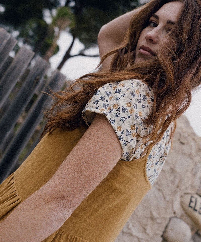 Joven mujer con vestido mostaza en un entorno natural, mirando pensativa sobre su hombro.